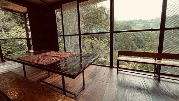 Een kamer met een glazen tafel, een bank en grote ramen met uitzicht op een bebost gebied.