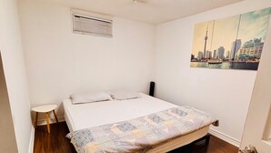 Une chambre à coucher avec un lit, une petite table de chevet, une bouche d’aération fixée au mur et un tableau représentant un paysage urbain.