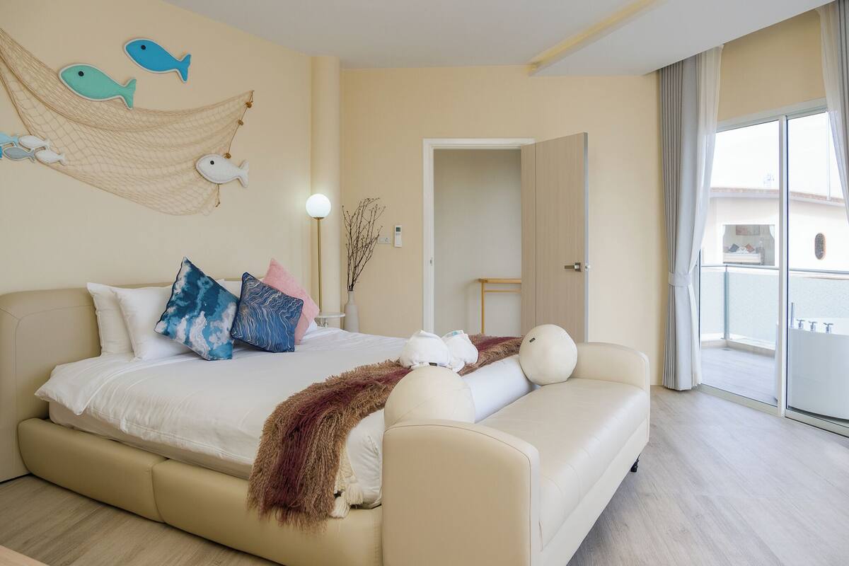 A bedroom with a bed, a sofa, a hammock with fish decorations, a lamp, and a view of the outside.