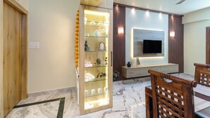 Een moderne hotelkamer met een houten deur, een glazen displaykast met objecten, een flatscreen televisie aan de muur en een eethoek met houten stoelen.
