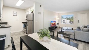 A modern kitchen with stainless steel appliances, a dining area with a table and chairs, a gray sofa, and a TV in the living room.