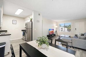 A modern kitchen with stainless steel appliances, a dining area with a table and chairs, a gray sofa, and a TV in the living room.
