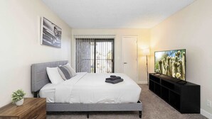 A bedroom with a bed, a TV, a window with blinds, a door, and a nightstand with a plant.