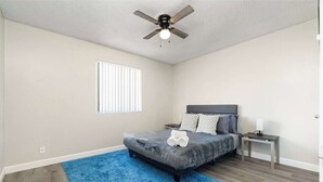 Un dormitorio con cama, ventilador de techo, ventana, mesita de noche y alfombra azul.