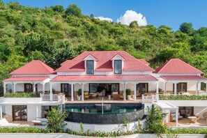 Una casa grande de varios pisos con piscina, varios balcones y un exuberante paisaje de colina de color verde.