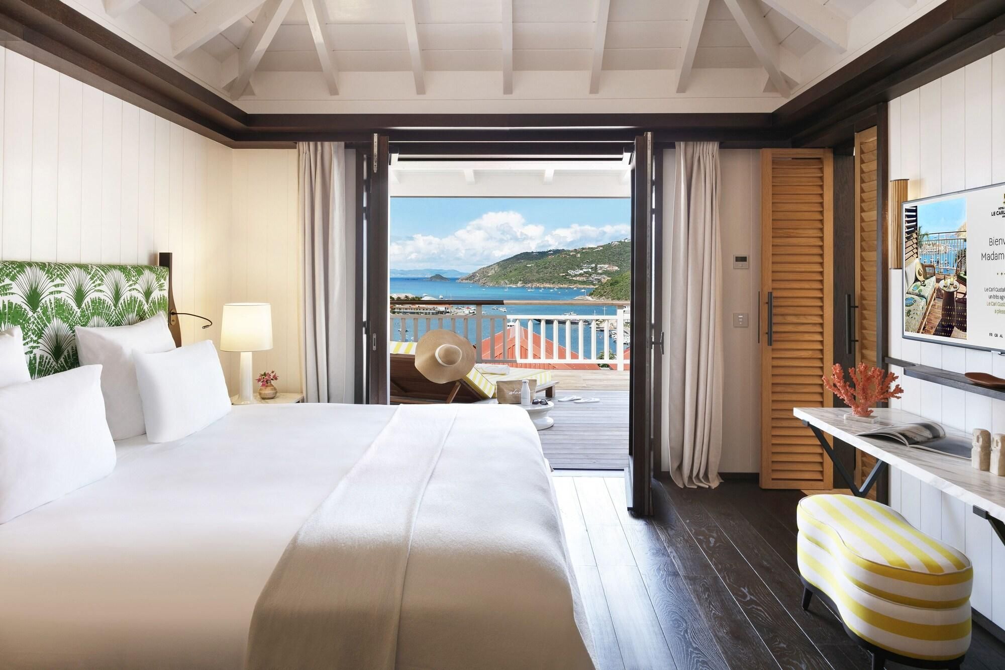 A hotel room with a bed, a desk, a chair, a TV, and a balcony with a view of the sea.