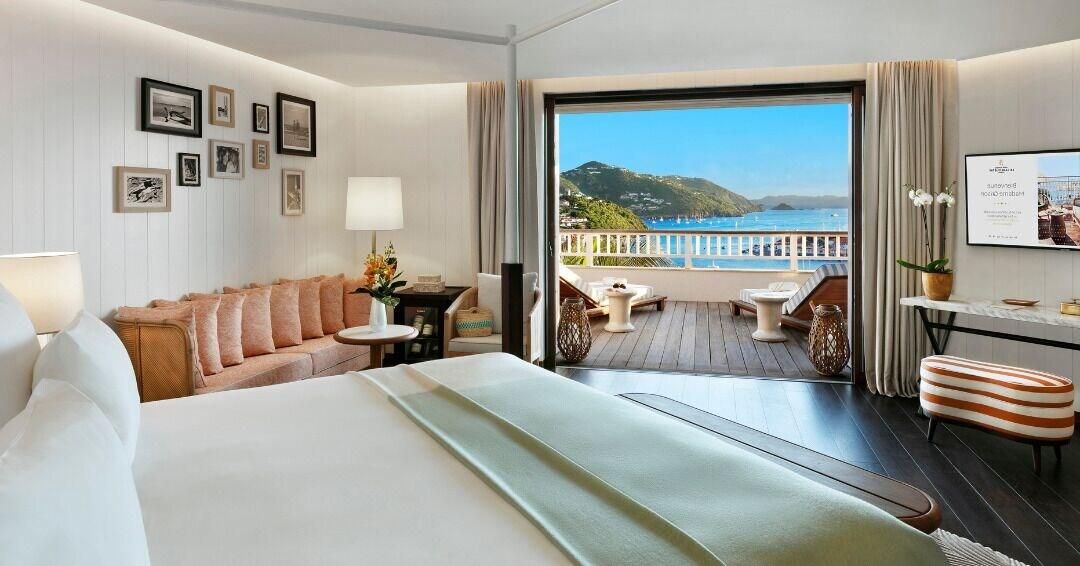 A hotel room with a bed, a sofa, a small table, a chair, and a balcony with a view of the sea.