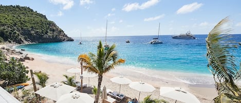 Uma praia com guarda-sóis, espreguiçadeiras e vista para o mar com barcos.