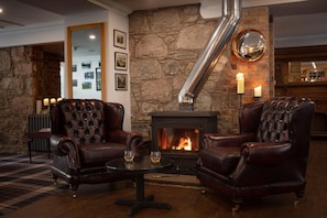 Une pièce avec un mur en pierre, une cheminée, des fauteuils en cuir, une table ronde et un miroir.