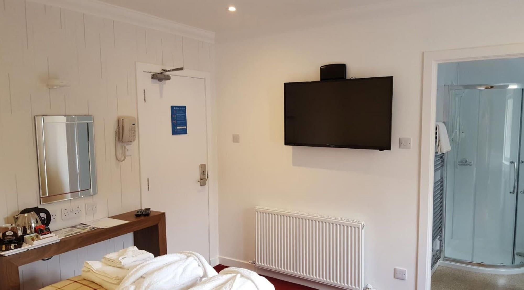 Habitación de hotel con cama, mesita de noche con tetera, espejo, detector de incendios, televisor, radiador y zona de ducha.