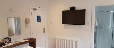 Habitación de hotel con cama, mesita de noche con tetera, espejo, detector de incendios, televisor, radiador y zona de ducha.
