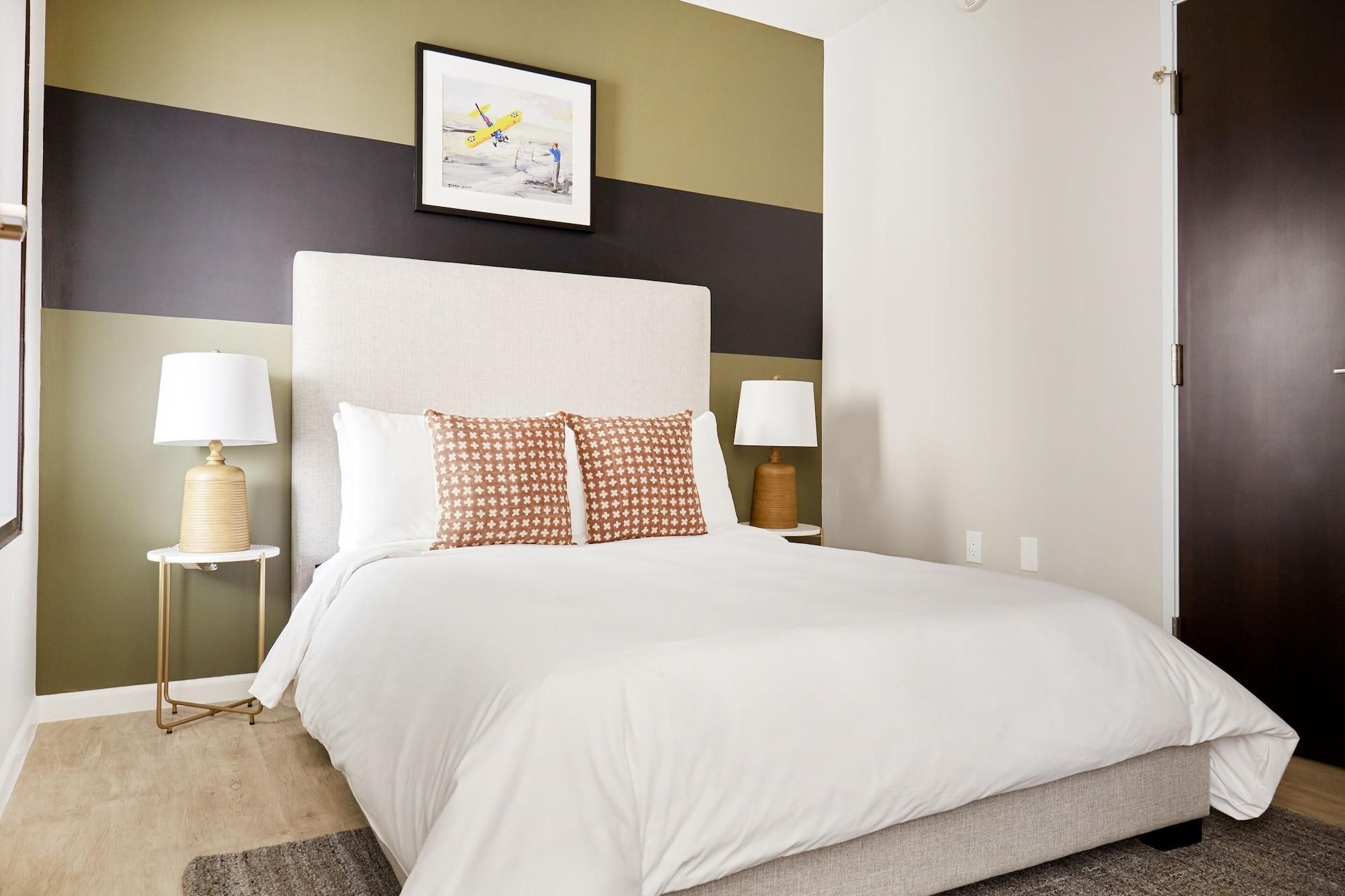 A hotel room with a bed, two bedside tables, a lamp on each table, a framed picture on the wall, and a door.