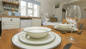 Een eettafel met een schaal en een wijnglas, een keuken op de achtergrond, en een raam met gordijnen.