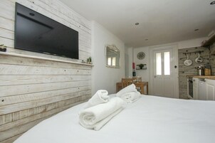 Un dormitorio con televisión montada en una pared de madera, una cama con toallas dobladas, una zona de comedor con sillas y una cocina con estufa y utensilios.