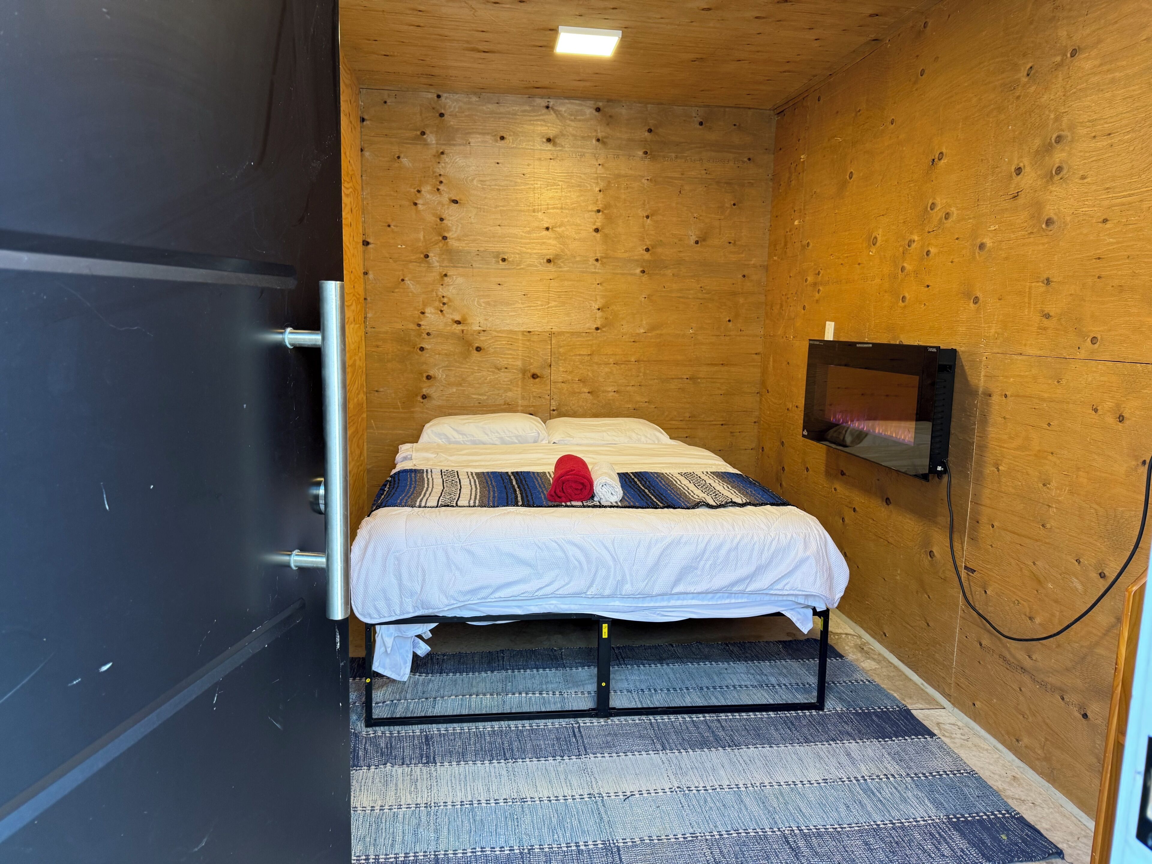 Een kleine, gezellige kamer met een houten wand, een bed met een geborduurde deken, een aan de muur bevestigde televisie en een bed met metalen frame.