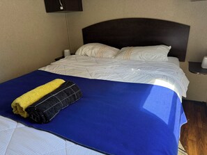 Habitació d'hotel amb un llit, una mantella blava, una tovallola groga i negra, almoines blanques i un capçal fosc.