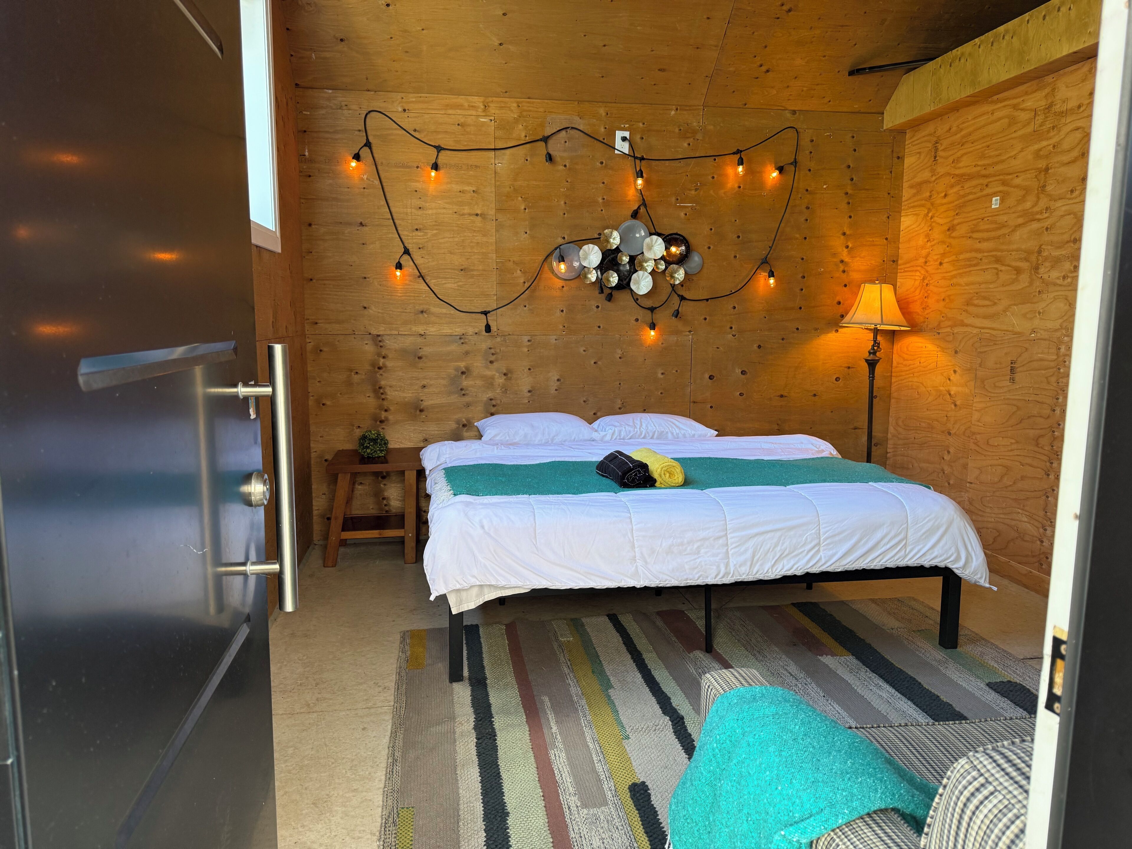 Un dormitorio con techo de madera, una cama con ropa blanca, una mesita de noche pequeña con una lámpara y una alfombra a rayas de colores.