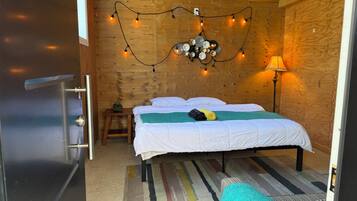 Un dormitorio con techo de madera, una cama con ropa blanca, una mesita de noche pequeña con una lámpara y una alfombra a rayas de colores.