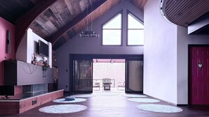 A spacious interior with a high ceiling, wooden beams, a fireplace, and a view through a glass door to an outdoor patio.