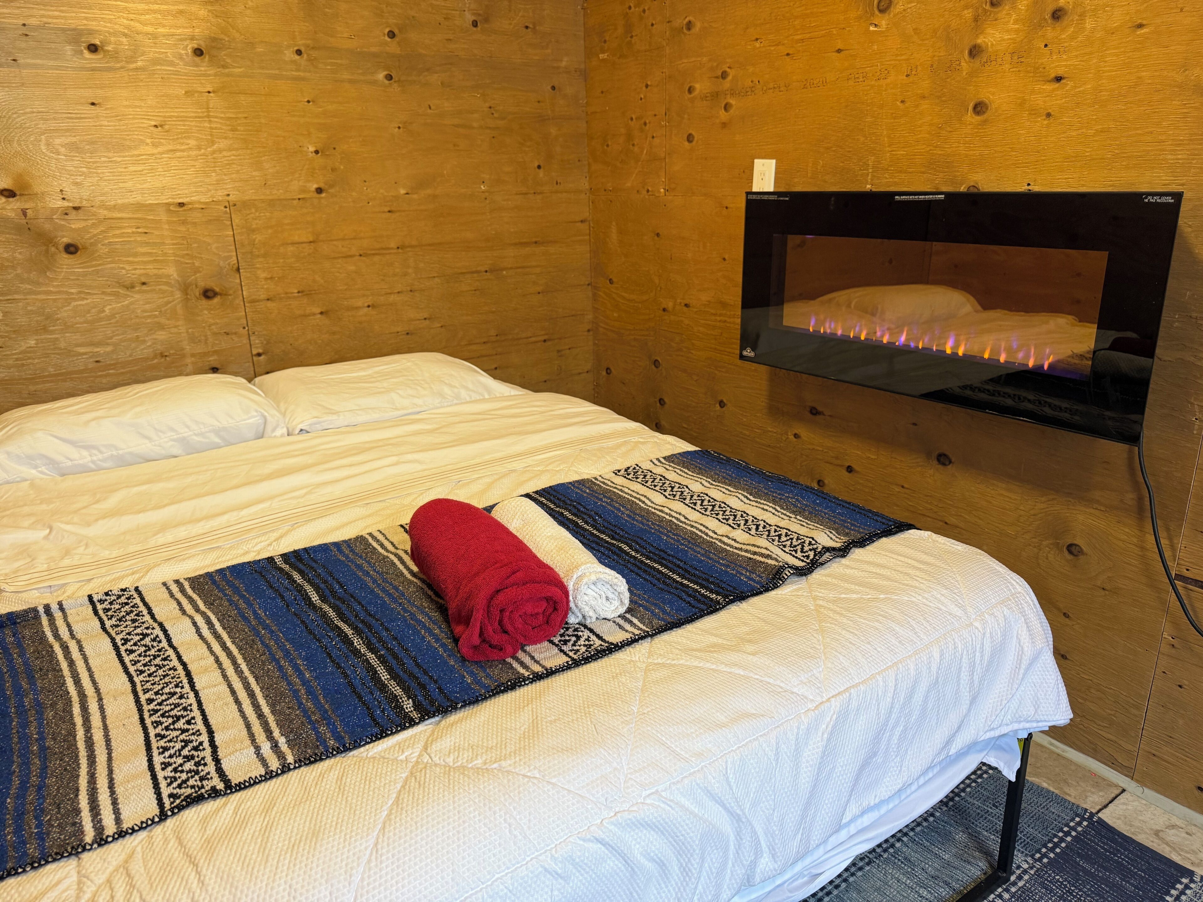 Una habitación de madera con una cama, un televisor montado en la pared y una manta con una toalla enrollada encima.