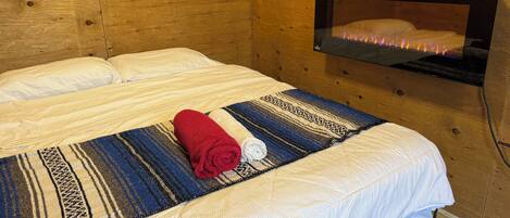 Una habitación de madera con una cama, un televisor montado en la pared y una manta con una toalla enrollada encima.