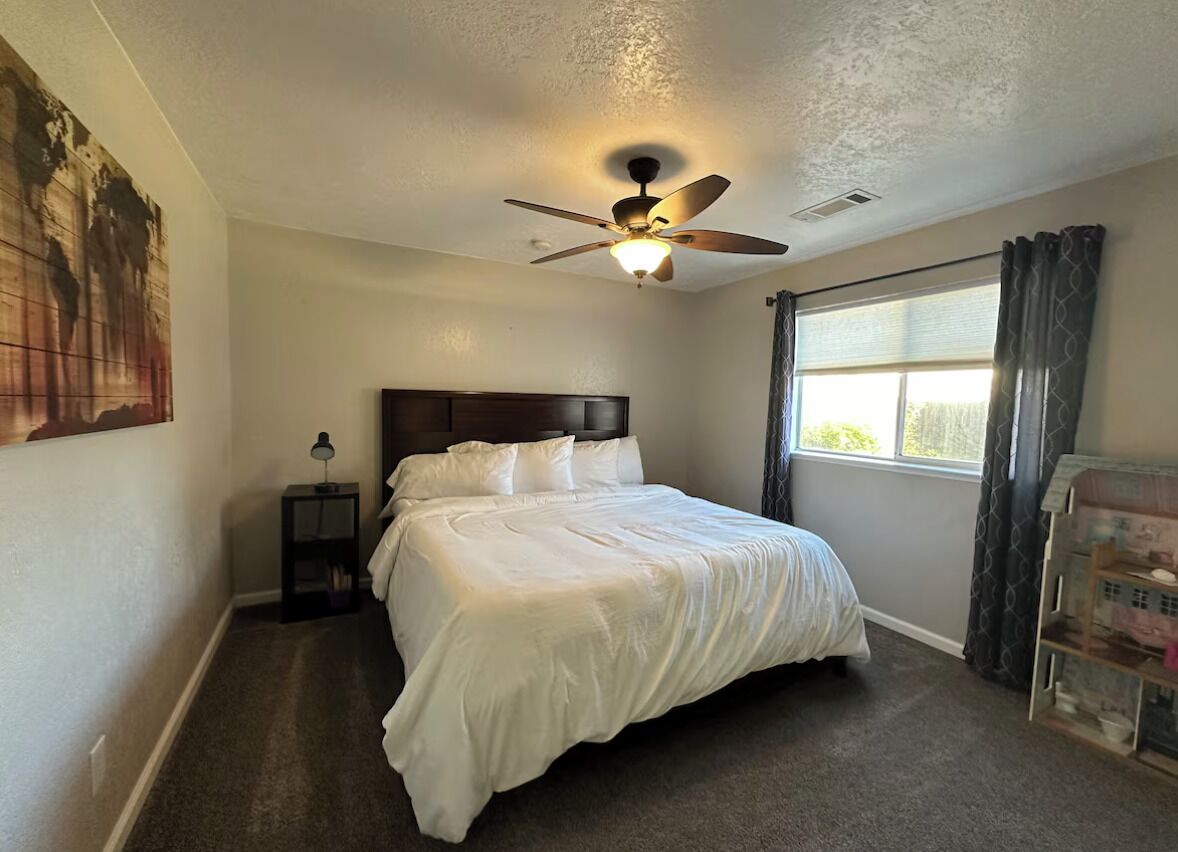 A bedroom with a bed, a ceiling fan, a nightstand, a lamp, a window with curtains, and a wall art piece.