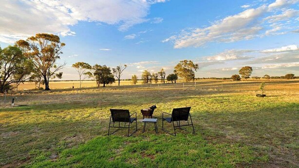 Un área de estar al aire libre con dos sillas y una mesa, rodeada por un campo cubierto de césped y árboles bajo un cielo despejado.