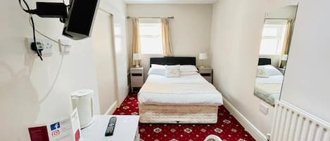 A hotel room with a bed, a desk with a computer, a chair, a TV mounted on the wall, and a window with curtains.