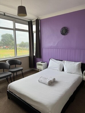 A hotel room with a bed, a nightstand, a chair, a window with curtains, and a clock on the wall. - Chatsworth Hotel (Skegness)