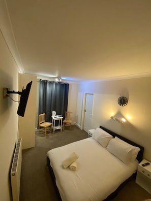 A hotel room with a bed, a desk, two chairs, a clock, and a TV mounted on the wall. - Chatsworth Hotel (Skegness)