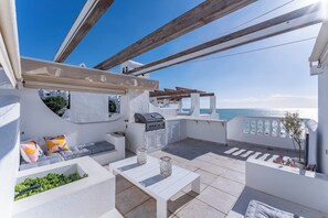 En udendørs terrasse med grill, siddepladser og havudsigt.