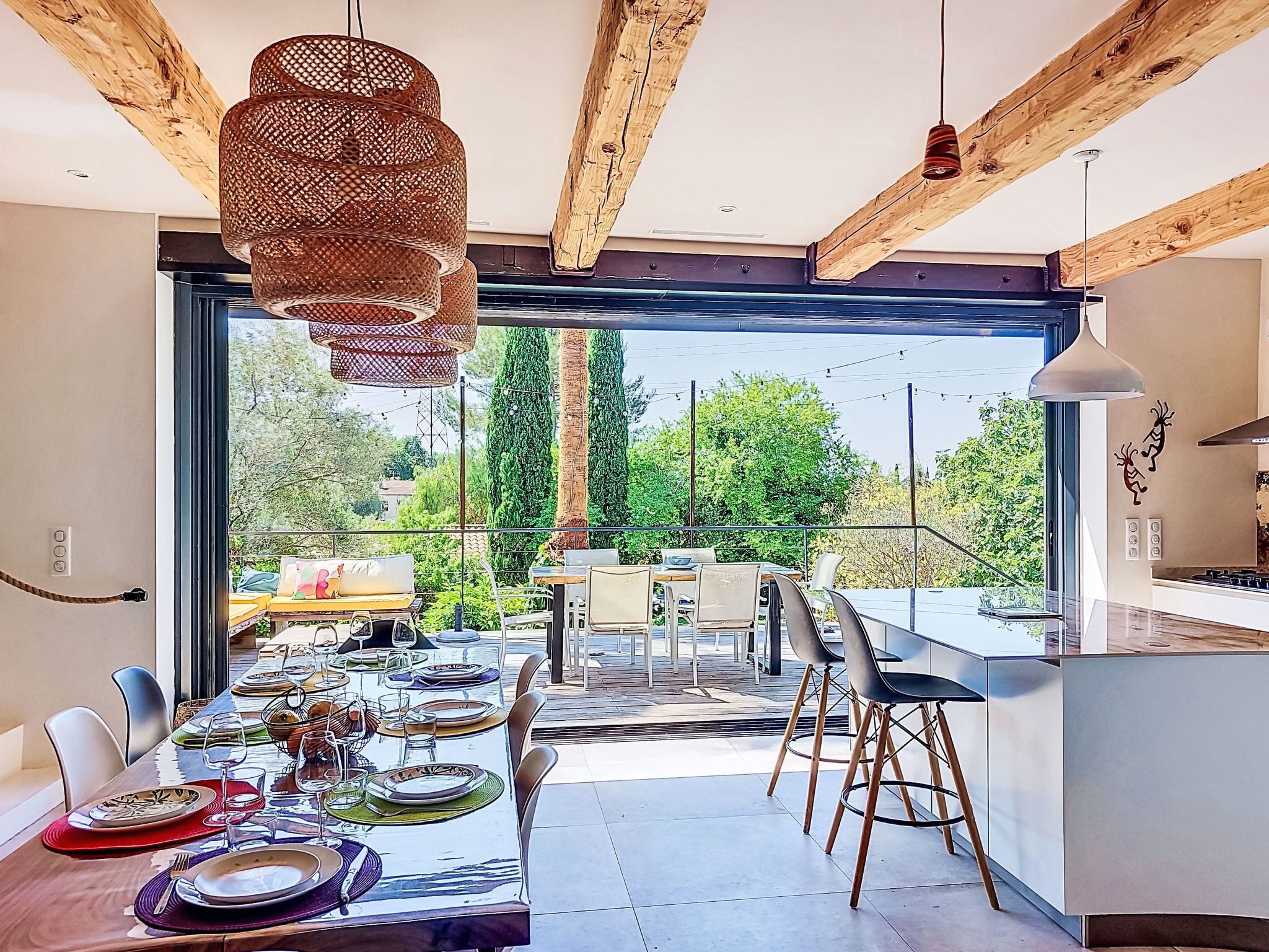 Un espace repas moderne avec une grande table dressée pour un repas, un coin cuisine avec des tabourets de bar et une vue sur la verdure à l’extérieur.