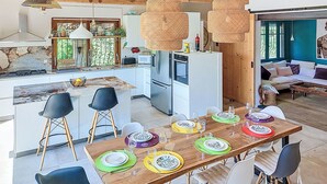 Ett modernt kök med en central köksö, ett matbord med plats för sex personer och ett vardagsrum med en soffa och ett stort fönster.