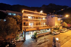 A multi-story hotel with a balcony on the ground floor, a signboard, and parked cars. - İsinda Pansiyon (Kaş)
