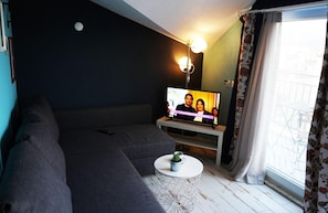 A living room with a sofa, a small round table with a plant, a TV on a stand, and a window with curtains. - İsinda Pansiyon (Kaş)