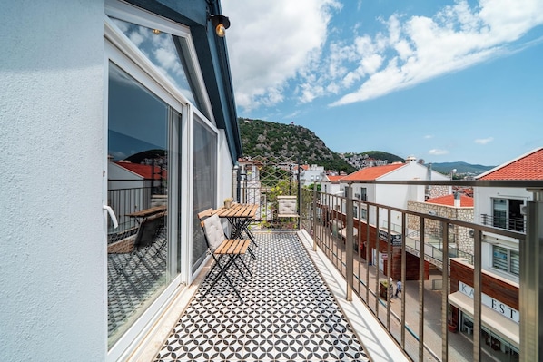 A balcony with a patterned floor, a table, and chairs, overlooking a street with buildings and a hill. - İsinda Pansiyon (Kaş)