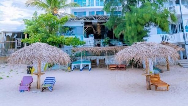 Un hotel frente al mar con una estructura de varios pisos, asientos exteriores con sombrillas de paja y una playa de arena.