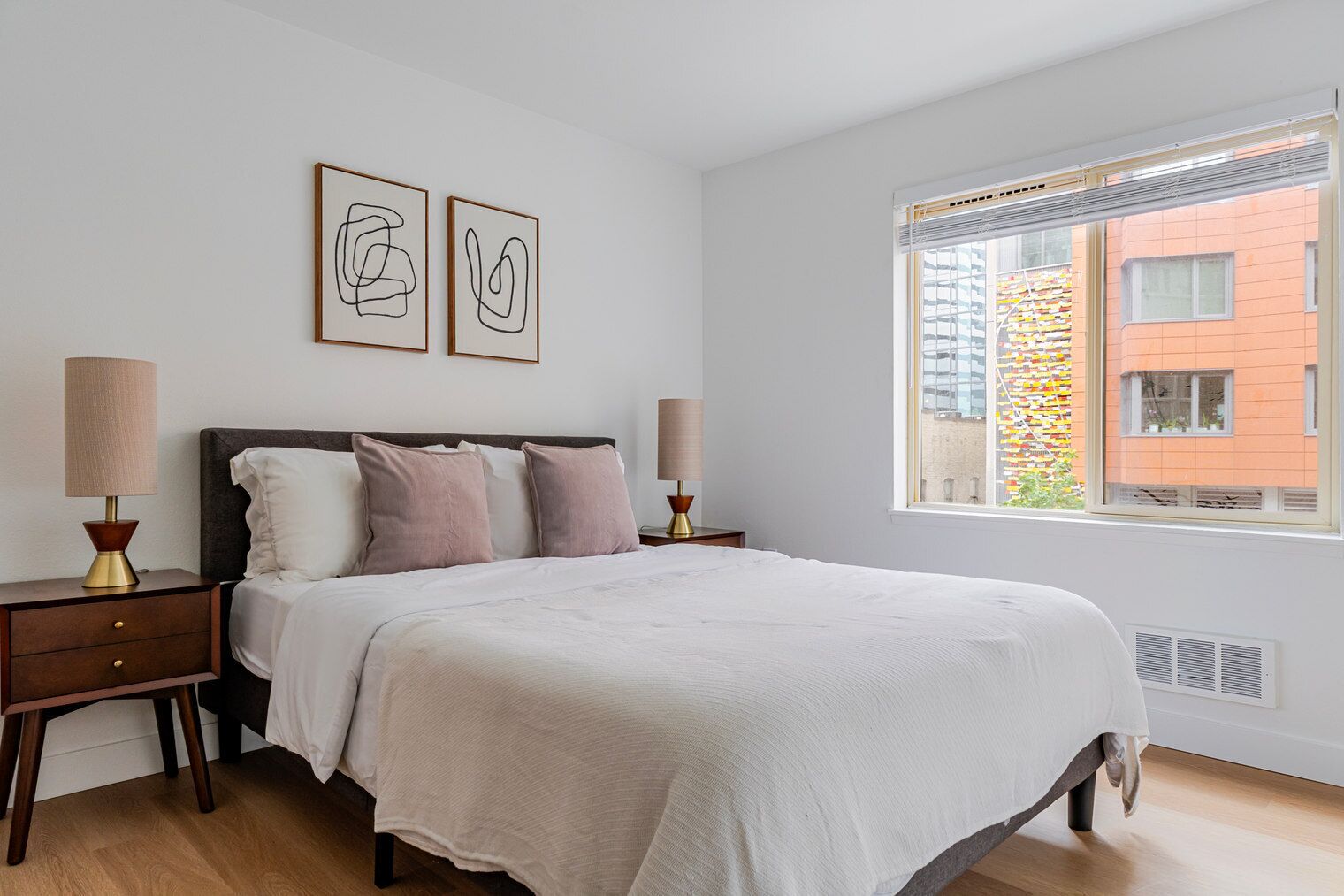 A bedroom with a bed, two bedside tables, two wall-mounted lamps, and a window with a view of buildings.