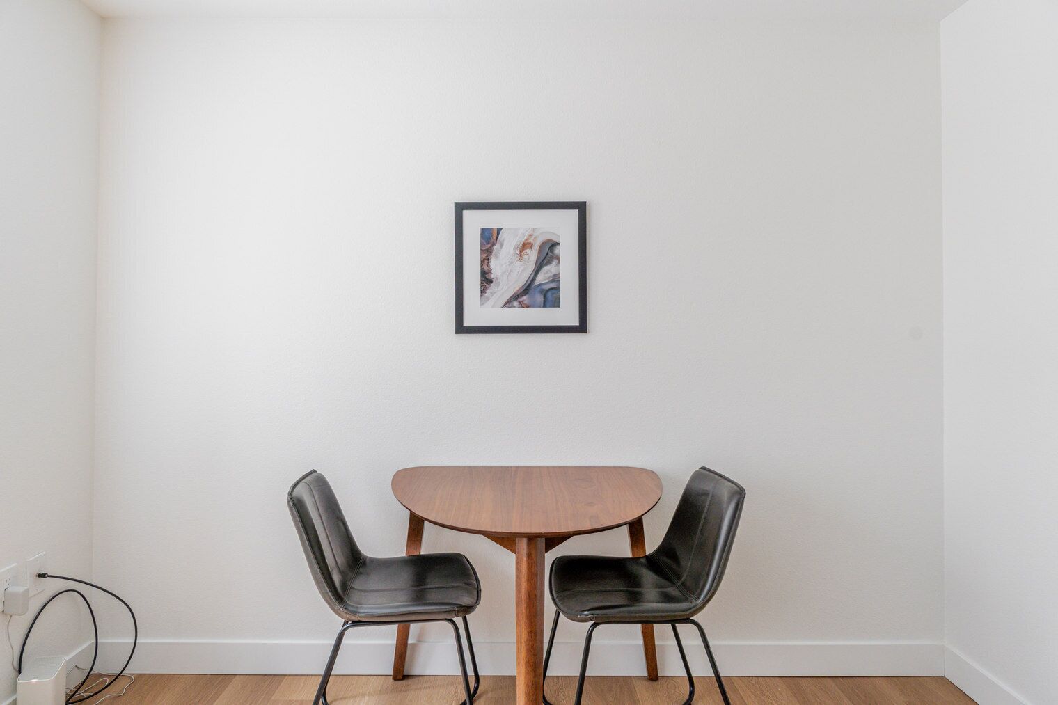 Un coin repas avec une table en bois et deux chaises noires, un tableau encadré au mur et un mur blanc.