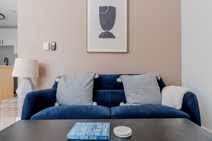 Ein modernes Wohnzimmer mit einem blauen Sofa, zwei Kissen, einem gerahmten Kunstwerk, einer Lampe und einem Buch auf einem Tisch.