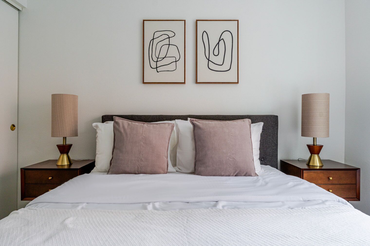 A neatly made bed with two pillows, two bedside tables with lamps, and two abstract art pieces on the wall.