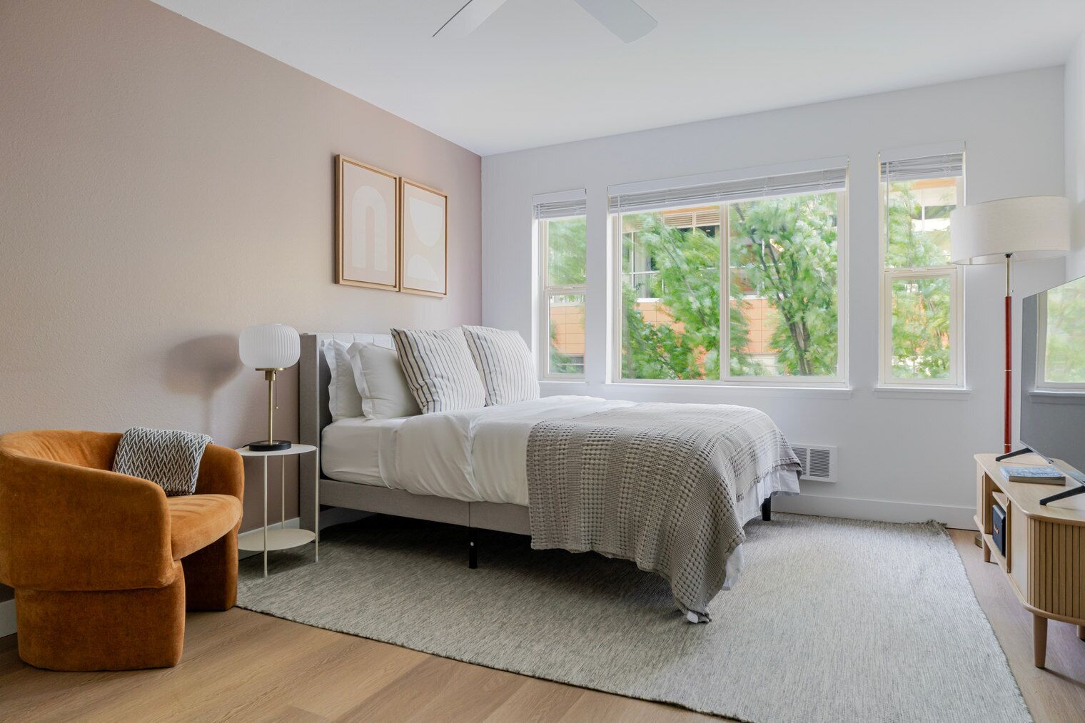 Quarto com cama, cadeira, mesa de cabeceira, luminária, televisão e janelas amplas com vista para o verde.