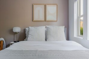 Ein ordentlich bezogenes Bett mit gestreiften Kissen, eine Nachttischlampe und zwei gerahmte abstrakte Kunstwerke an der Wand.