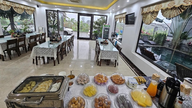 A dining area with tables, chairs, a buffet with various food items, and a large window with a view of greenery. - Lotus Hotel (Nam Hoa Lu)