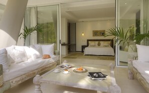 A modern hotel room with a glass door leading to a balcony, a bed with white bedding, a coffee table with a magazine and croissant, and a sofa with white cushions.