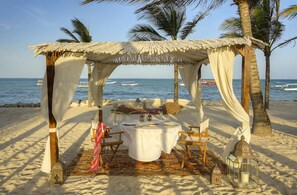 Una cabaña de playa con una mesa dispuesta para dos, rodeada de palmeras y con vistas al mar.