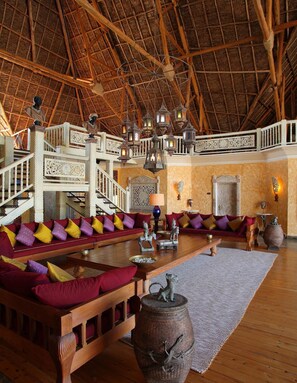 A spacious lounge with a high wooden ceiling, ornate railings, and a central seating area with cushions.