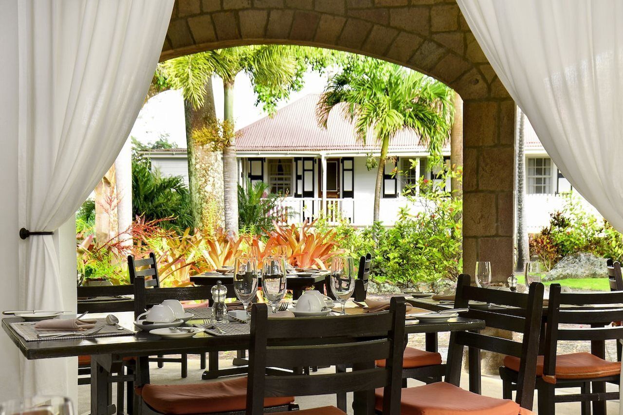 An outdoor dining area with tables set for two, surrounded by greenery and a stone archway.