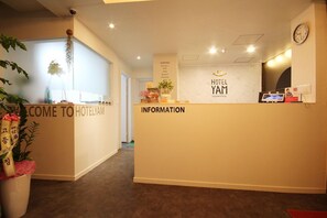 Zona de recepción de hotel con mostrador, un reloj y un cartel con las etiquetas 'HOTEL YAM' e 'INFORMACIÓN'.