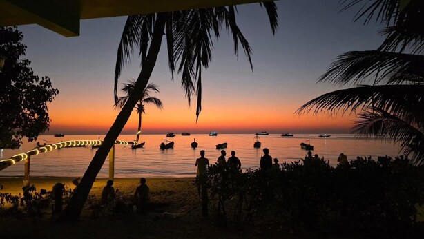 Una playa al atardecer con barcos en el agua y gente al frente.